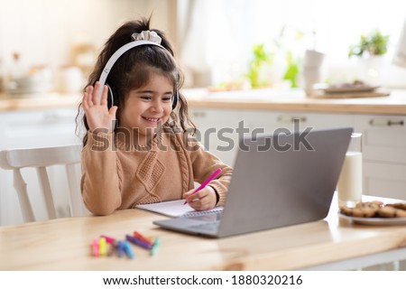 Distance Learning. Cute little home schooler girl having video lesson with laptop in kitchen, studying with computer during coronavirus quarantine, enjoying online education, waving hand at camera