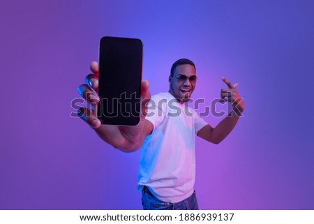 Cool Mobile Offer. Excited African Guy Demonstrating Smartphone With Black Screen, Standing Under Neon Lighting, Joyful Millennial Man Showing Free Mockup Copy Space For Your App Or Website Design