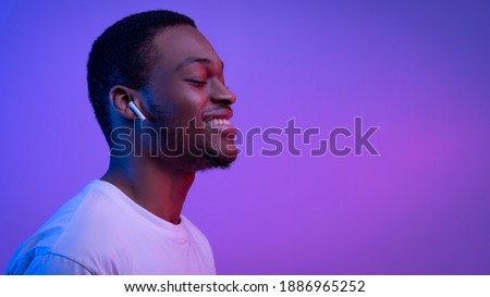 Relaxed African American Man Listening Music In Wireless Airpods Earphones In Neon Light, Smiling Black Guy Wearing Bluetooth Earpods Enjoying Favorite Songs Over Purple Background, Panorama