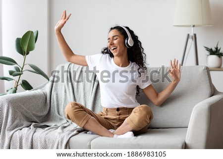 Carefree brunette woman listening to music and singing, using headphones, sitting on couch in living room, copy space. Happy young lady with closed eyes enjoying music, home interior