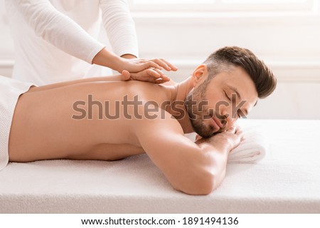 Side view of relaxed handsome man having body massage at spa salon. Middle aged bearded man attending modern male spa, laying on massage table, getting healing body procedure, enjoying his day at spa