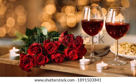 Romantic Dinner. Bouquet of flowers lying on the table, selective focus on bunch of roses, two glasses of red wine and candles on the wooden desk. Date concept, blurred background, banner, copy space