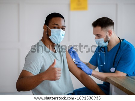 African American man in antiviral mask gesturing thumb up during coronavirus vaccination, approving of covid-19 immunization. Young doctor giving vaccine injection to male patient