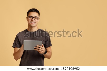 Modern work, social media and online communication. Attractive happy young male student in glasses, with smart watch typing in digital tablet, isolated on beige background, studio shot, free space