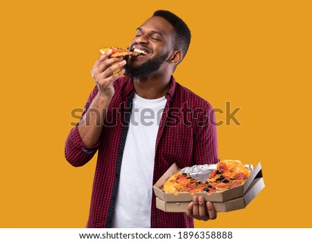 Happy African Young Man Enjoying Pizza Biting Tasty Slice Posing With Box Over Yellow Background. Junk Food Lover Eating Italian Pizza In Studio. Unhealthy Male Nutrition And Cheat Meal