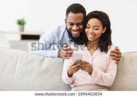 Smiling young black man embracing woman from behind while looking at smartphone. African american couple sharing social media on mobile phone, happy female sitting on the couch, showing gadget