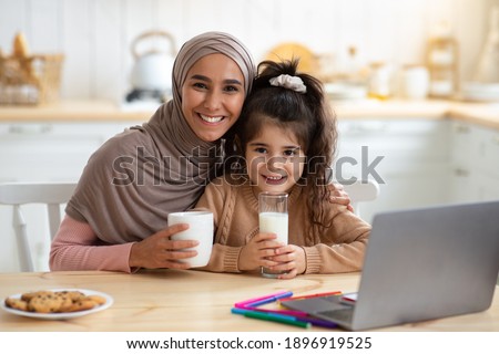 Happy Muslim Mom And Daughter Having Snacks And Drinks In Kitchen, Happy Islamic Lady In Hijab Enjoying Coffee And Her Cute Little Child Drinking Milk. Cheerful Family Smiling At Camera, Free Space