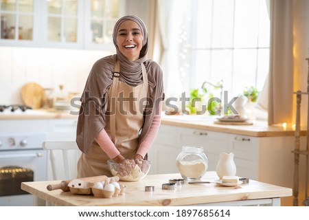 Baking Concept. Portrait Of Joyful Muslim Woman In Hijab Kneading Dough In Kitchen Interior, Cheerful Islamic Female In Hijab And Apron Having Fun While Preparing Homemade Pastry, Laughing At Camera
