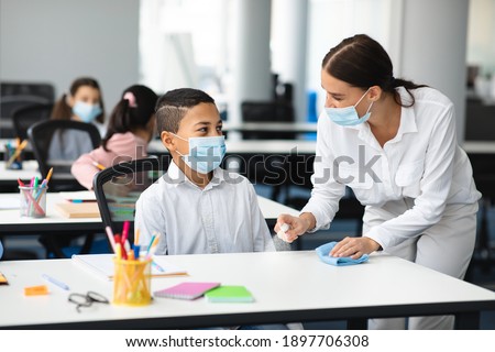 Hygiene And Cleaning. Teacher cleaning table with antibacterial sanitizer and napkin, disinfecting surface, applying antiseptic spray on desk, wearing disposable face mask, boy studying in classroom