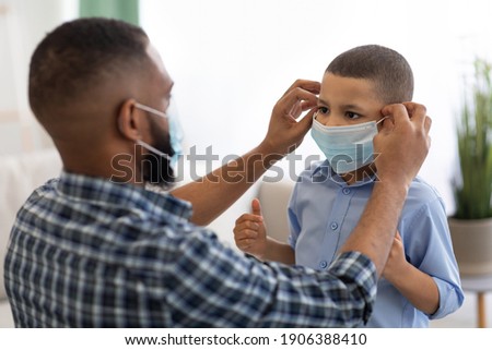 Coronavirus Prevention. Black Dad Putting On Protective Surgical Mask On Son's Face Before Walk Standing At Home. Covid-19 Protection And Healthcare Concept. Selective Focus ストックフォト © 
