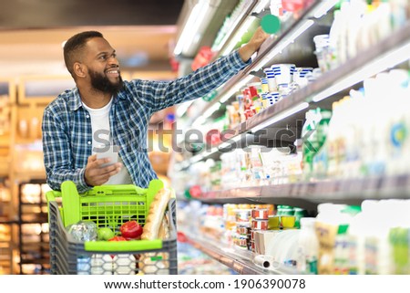 Black Male Buyer Shopping Groceries In Supermarket Taking Dairy Product From Shelf Standing With Shop Cart Indoors. Guy Buys Grocery Choosing Food In Super Market. Empty Space