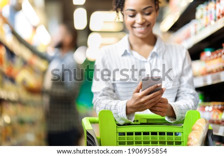 African Lady Customer Doing Grocery Shopping Using Smartphone Walking With Cart In Supermarket. Selective Focus. Woman Using Groceries Shopping Application On Phone Bying Food In Super Market