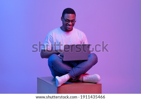 Handsome Black Programmer Guy Using Laptop Computer, Sitting On Big Cube In Neon Lighting, Cheerful African American Man In Glasses Working Or Study Online Over Purple Studio Background, Copy Space