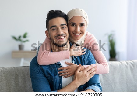 Portrait of cheerful muslim couple embracing at home, copy space, closeup. Smiling young woman in headscarf hugging her loving husband from behind, middle-eastern family spending time together