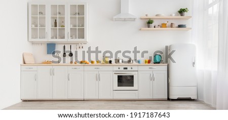 Interior of modern comfortable kitchen. Multicolored cups and teapot, orange juice in glass and utensils on white furniture, refrigerator, flowers in pots on shelves, light wall in daylight, panorama