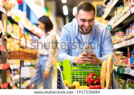 Smiling Male Customer Doing Grocery Shopping Using Smartphone Walking With Cart In Supermarket. Selective Focus. Man Using Groceries Shopping Application On Phone Bying Food In Super Market