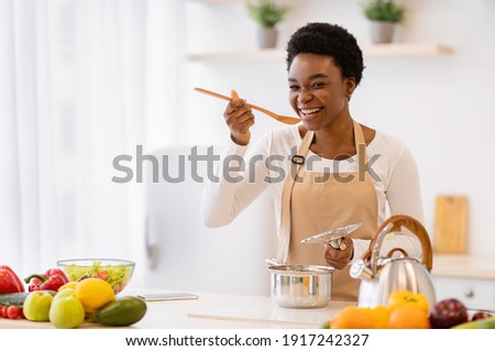 Happy African American Woman Cooking Tasting Dinner In A Pot Standing In Modern Kitchen At Home. Housewife Preparing Healthy Food Smiling To Camera. Household And Nutrition. Dieting Recipes Concept