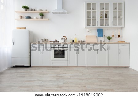 Light kitchen in daylight, simply, minimalist scandinavian interior. White furniture, small refrigerator, stove with utensils, extractor hood, shelves with plants in pots, curtains in morning