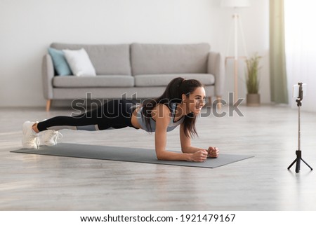 Joyful young lady in sportswear planking in living room, watching fitness video or having video conference with fitness trainer, sporty woman exercising and watching at smartphone screen, free space