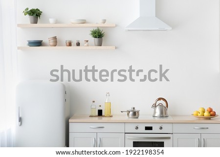 Stylish modern apartment with minimalist kitchen and Scandinavian interior. White furniture with stove, kettle and utensils. Fruits in plate, shelf with dishes and plants in pots, light wall