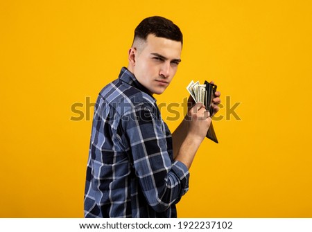 Young man with wallet full of money being greedy over his wealth on orange studio background. Millennial guy with dollars acting mean and stingy, not willing to pay taxes or share his income Photo stock © 