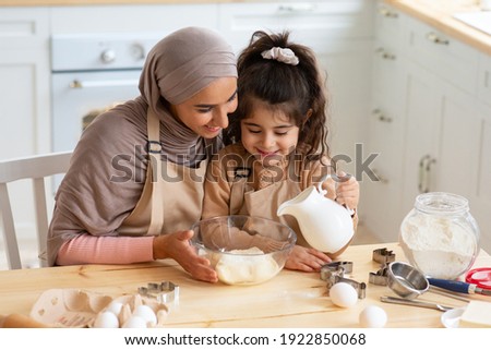 Young Muslim Mom Baking Together With Her Little Daughter In Kitchen. Cute Little Arab Girl Helping Mom To Prepare Dough, Adding Ingredients To Bowl, Islamic Family Having Fun While Cooking At Home