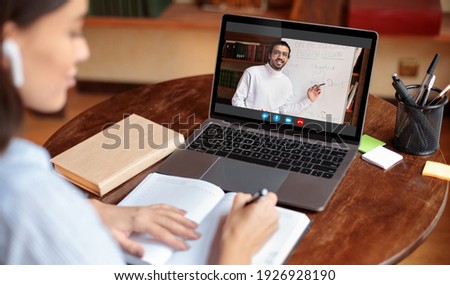 Distant Education Concept. Over the shoulder view of young woman wearing earbuds sitting at table and writing in her notebook, taking notes and using laptop, having online class