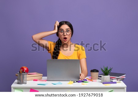 Wow. Portrait of shocked amazed indian lady in eyeglasses sitting at table with laptop and grabbing her head isolated on purple studio background. Stressed young woman can't believe news Stock foto © 
