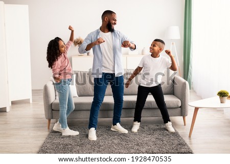 Spending Time Together Concept. Cheerful black dad moving and dancing to music with his excited daughter and son. Playful children having fun with father, enjoying weekend with family in living room