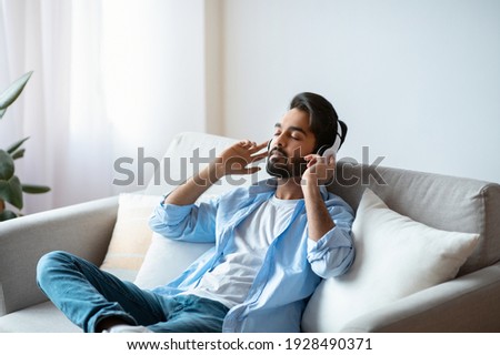 Weekend Rest. Young arab guy in wireless headphones listening audiobook at home, calm eastern man sitting with closed eyes on couch in living room, enjoying domestic leisure, free space Foto stock © 