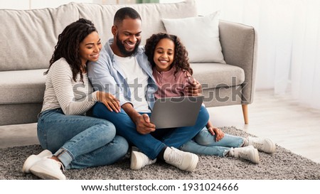 Family Weekend. Cheerful African American parents and their little daughter using laptop at home together, watching movie or browsing internet, sitting on the floor carpet in living room