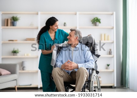 Young nurse covering disabled elderly man in wheelchair with warm plaid at retirement home. Millennial caregiver assisting handicapped senior patient, taking care of older male indoors
