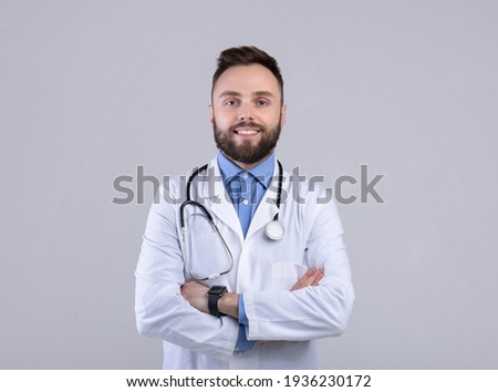 Portrait of positive male doctor posing with crossed arms and smiling at camera over grey studio background