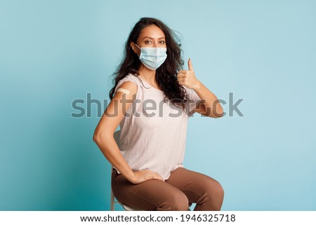 Covid-19 Vaccination. Vaccinated Woman Gesturing Thumbs Up Showing Arm With Plaster Bandage After Coronavirus Vaccine Injection On Blue Background, Wearing Protective Face Mask. Antivital Immunization