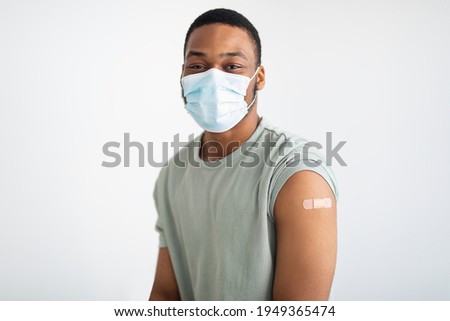 Coronavirus Vaccination. Portrait Of Vaccinated African Guy Wearing Protective Face Mask Looking At Camera Posing With Adhesive Tape On Arm After Vaccine Injection Over White Background
