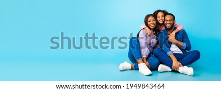 Happy Loving Family. Black man, woman and girl sitting on the floor isolated on blue studio wall. Smiling daughter emracing her mum and dad from behind, posing at camera, banner, panorama, copy space 商業照片 © 
