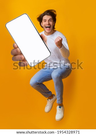 New awesome mobile app. Jumping emotional young guy showing smartphone with blank screen on orange studio background, mockup for application or website design