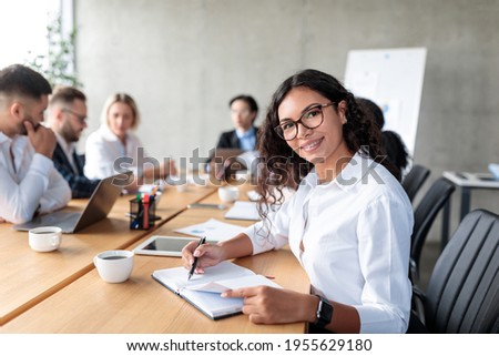Mexican Businesswoman Posing Taking Notes And Smiling To Camera Sitting At Corporate Business Meeting With Coworkers In Office. Female Entrepreneurship Career Concept