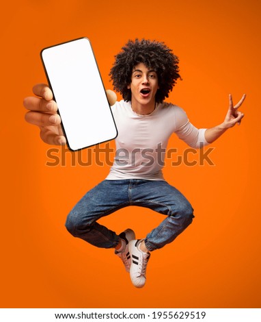 Portraif of excited African American teen guy jumping with smartphone, demonstrating empty screen on orange background, copy space for your mobile advertisement, mockup image