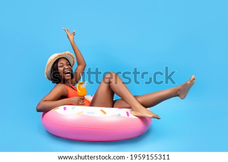 Positive black woman sitting on inflatable ring, holding tropical cocktail on blue background, full length. Happy African American lady having beach party with pool float and refreshing summer drink