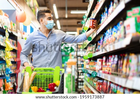 Eastern Arabic Guy Buying Food Doing Grocery Shopping Walking With Shop Cart Full Of Groceries In Modern Supermarket, Wearing Face Mask. Male Customer Choosing Staple Products On Shelf In Store Foto stock © 