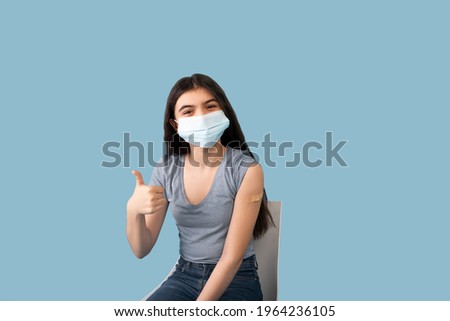 Vaccinated teen girl in face mask showing plaster bandage on her arm after getting covid-19 vaccine injection on blue studio background. Coronavirus population immunization campaign