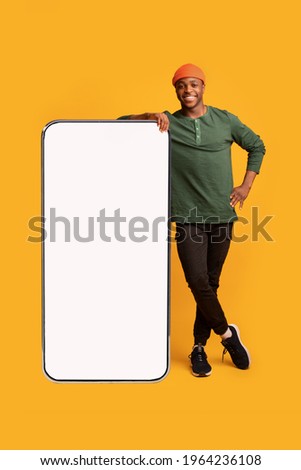 Mobile Offer. Happy Black Guy Leaning At Big Smartphone With Blank White Screen, Cheerful African Man Demonstrating Copy Space For Your Design Or Advertisement, Posing Over Yellow Background, Mockup