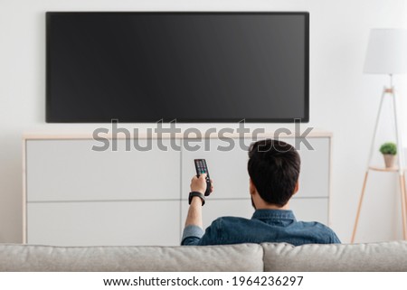 Man watching television, turning on plasma flatscreen TV-set, pointing remote control at empty TV screen on white wall. Guy switching channels at home, back view. Mockup Photo stock © 