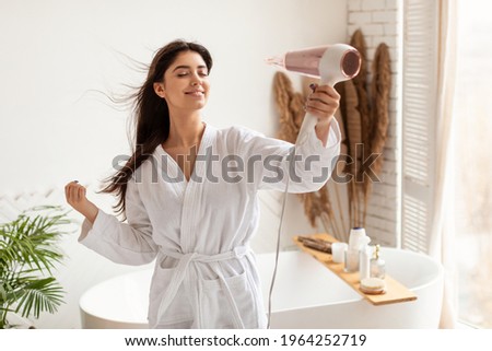 Haircare. Brunette Woman Drying And Styling Hair With Hairdryer Making Hairstyle Standing In Modern Bathroom At Home. Happy Female Using Hair Dryer Enjoying Beauty Routine After Morning Shower