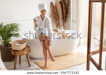 Woman In Bathrobe Moisturizing Smooth Legs With Moisturizer Cream Sitting On Bathtub In Modern Bathroom Indoors After Morning Shower. Beauty, Skincare And Pampering Concept
