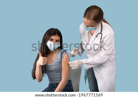 Successful covid-19 vaccination concept. Teenage girl in face mask showing thumb up gesture while being immunized against coronavirus during her visit to doctor, blue studio background