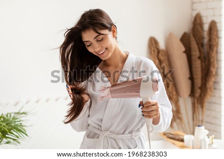 Young Woman Drying And Styling Hair Using Hairdryer Standing In Bathroom At Home After Morning Shower. Haircare And Hairstyling Products And Devices Concept. Female Beauty Routine Concept