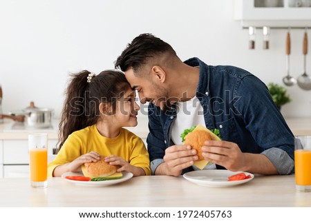 Happy Loving Arab Dad And Daughter Eating Sandwiches In Kitchen And Bonding, Cheerful Middle Eastern Father And Cute Little Female Child Touching Foreheads And Smiling, Enjoying Time Together