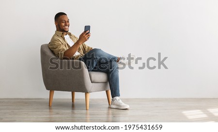 Cheerful African American Guy Using Mobile Phone With New Application Sitting In Armchair Over Gray Wall Background. Gadgets And Mobile Communication Concept. Panorama, Empty Space For Text